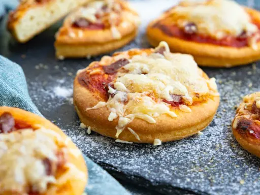 Mehrere mini pizza mit geschmolzenem Käse und Tomatensauce auf einem dunklen Untergrund, bestreut mit etwas Mehl.