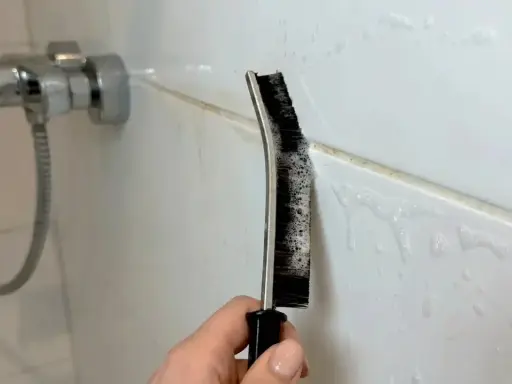 Hand reinigt Duschfugen mit einer Bürste und Schaum.