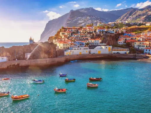 Bunte Häuser schmiegen sich an steile Hänge auf den Inseln bei Madeira. Kleine Boote schaukeln im azurblauen Wasser vor einem Leuchtturm.