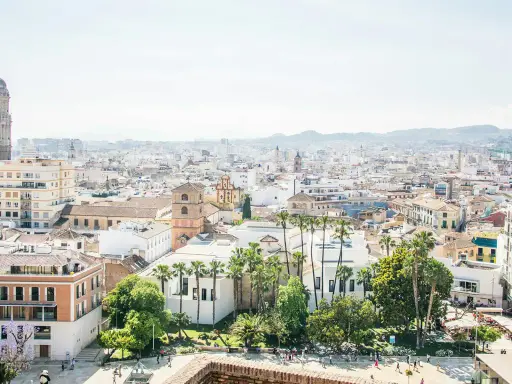Blick über die Dächer von Malaga, mit dem markanten Glockenturm der Kathedrale und Palmen. Der malaga hafen ist in der Ferne zu erahnen.