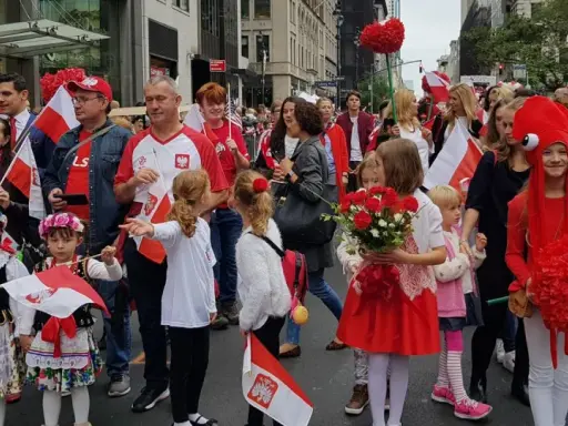 Dzieci i dorośli w strojach ludowych i z polskimi flagami maszerują ulicą. Widać serca z czerwonych balonów i kwiatów. Polacy w Szwecji świętują.