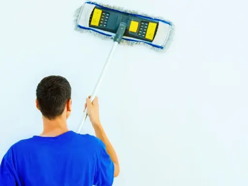 Hombre con camiseta azul limpiando paredes blancas con un plumero.