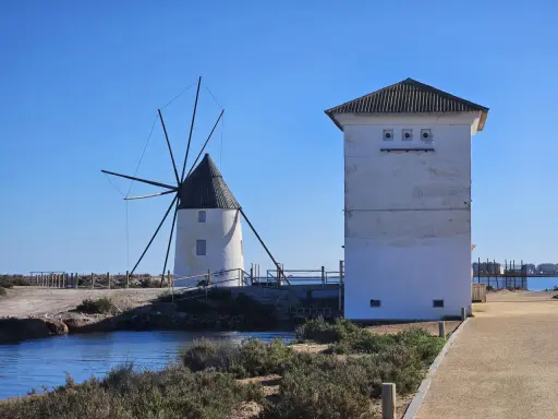 Altes Windrad und ein weißes Gebäude am Mar Menor, mit Wasser und Vegetation im Vordergrund.