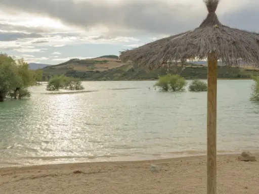 Sombrilla de paja en la orilla del pantano de Alloz. El agua turquesa, árboles emergiendo y colinas al fondo invitan a descubrir cómo llegar.