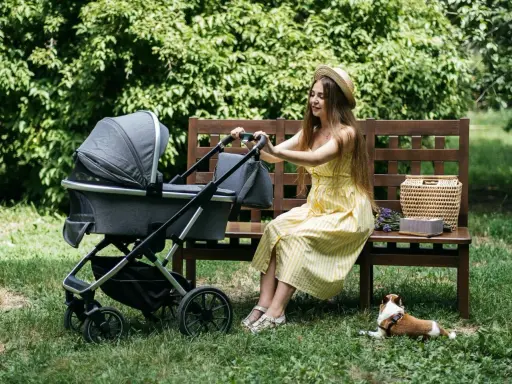 Kobieta w żółtej sukience siedzi na ławce obok wózka Kinderkraft. Piesek leży na trawie.