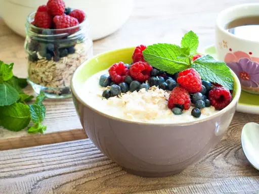 Schüssel mit Haferflocken mit Kefir, garniert mit Himbeeren, Blaubeeren und Minze. Daneben ein Glas Müsli und eine Tasse Tee. Gesundes Frühstück.