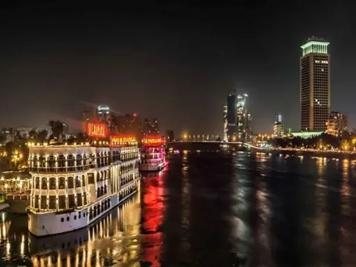 Beleuchtete Nilschiffe und moderne Hochhäuser spiegeln sich im Wasser. Ein magischer Anblick von Kairo bei Nacht.