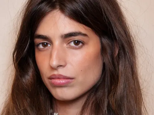 Portrait d'une jeune femme aux longs cheveux bruns. Son maquillage met en valeur ses yeux et ses lèvres, avec un soin particulier apporté à son sourcil poil à poil.