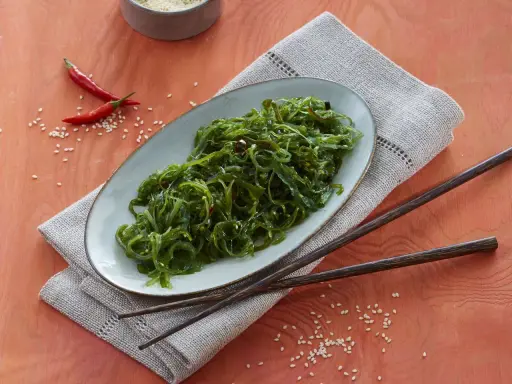 Ein Teller mit leckerem Wakame Salat, garniert mit Chilis und Sesam. Dazu liegen Holzstäbchen bereit.