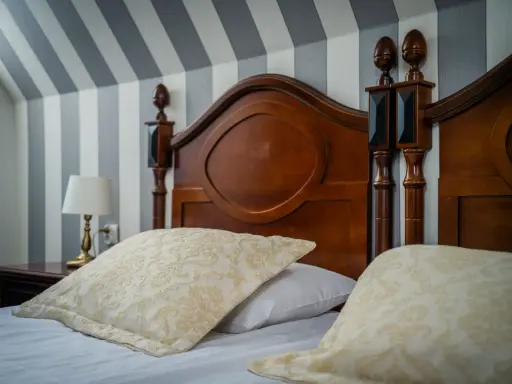 Eleganckie łóżko z drewnianym zagłówkiem i ozdobnymi poduszkami. Ciekawe, ile wynosi ryczałt za nocleg w takim miejscu?
