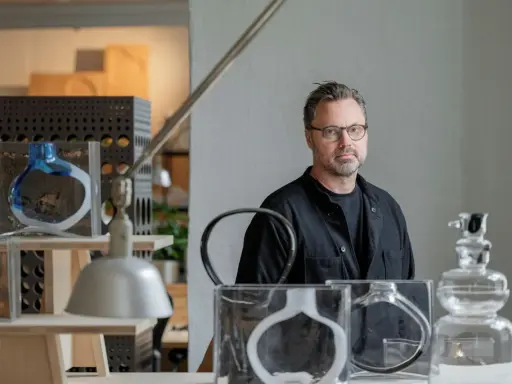 Matti Klenell, en man med glasögon, står bland sina konstverk av glas.