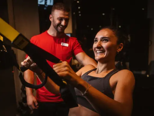 Frau lacht und trainiert mit TRX-Bändern, ihr Trainer lächelt. Sie will schnell wieder fit werden.