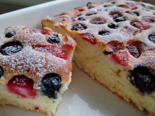 Puszyste ciasto na maślance z jagodami i malinami, posypane cukrem pudrem. Idealne na deser.