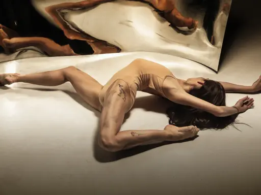 Młoda kobieta w cielistym body wygina się w łuk na podłodze, jej odbicie w lustrze tworzy surrealistyczny obraz. Jak z opowiadanie erotyczne nastolatki.