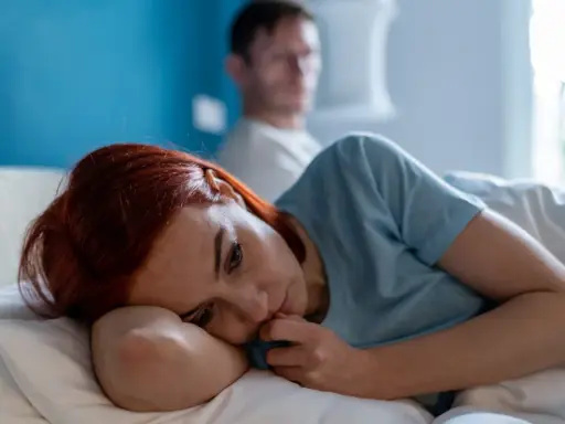 Zmartwiona kobieta leży w łóżku, podczas gdy jej partner jest niewyraźny w tle. Scena sugeruje **obsesyjne kontrolowanie partnera** i jego skutki.
