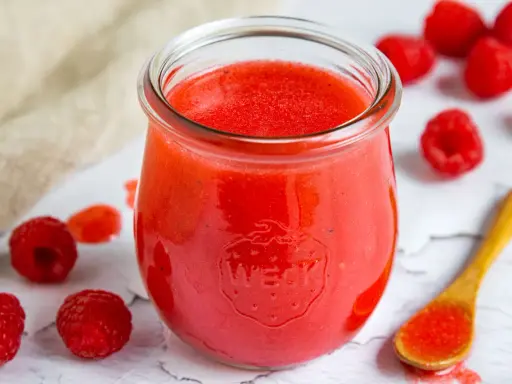 Ein Glas mit leuchtend roter Himbeer-Vinaigrette, umgeben von frischen Himbeeren und einem Holzlöffel mit etwas Vinaigrette.