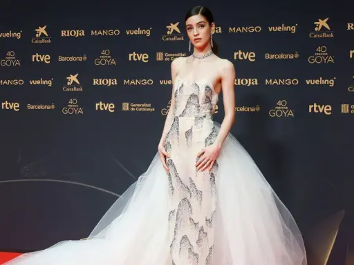 Una joven desfila en la goya alfombra roja con un vestido de gala blanco con detalles plateados y una voluminosa cola de tul.
