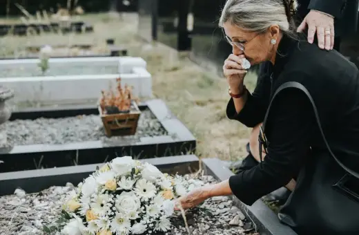 Kobieta w żałobie klęczy przy grobie, ocierając łzy. Czy zasiłek pogrzebowy należy się każdemu? Wieniec z białych i żółtych kwiatów leży na ziemi.