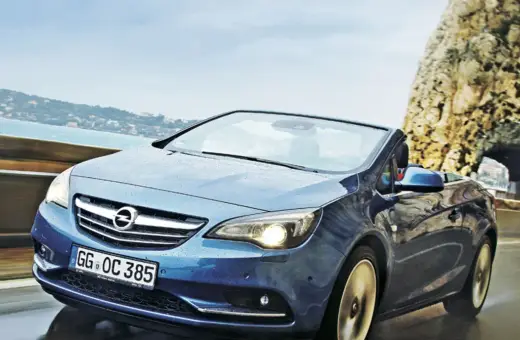 Niebieski Opel Cascada z opuszczonym dachem jedzie po mokrej drodze przy skalistym wybrzeżu.