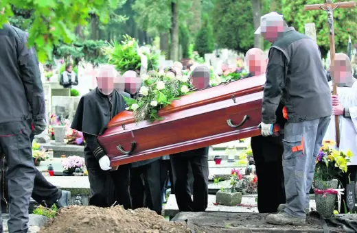 Pogrzeb na cmentarzu. Kilku mężczyzn niesie drewnianą trumnę ozdobioną kwiatami w kierunku grobu głębinowego.