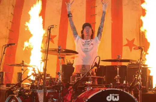Tommy Lee w ogniu, z rękami w górze i pałeczkami perkusyjnymi, na tle płonącej sceny.