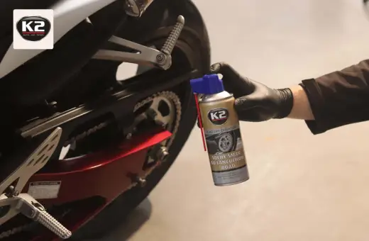 Ręka w czarnej rękawiczce trzyma puszkę smaru do łańcucha motocyklowego K2 przy tylnym kole motocykla.