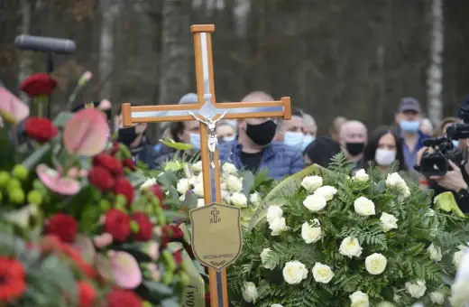 Drewniany krzyż z figurą Chrystusa na pogrzebie. Wieniec białych róż i czerwonych kwiatów. Ludzie w maseczkach. Co zrobić z krzyżem po pogrzebie?