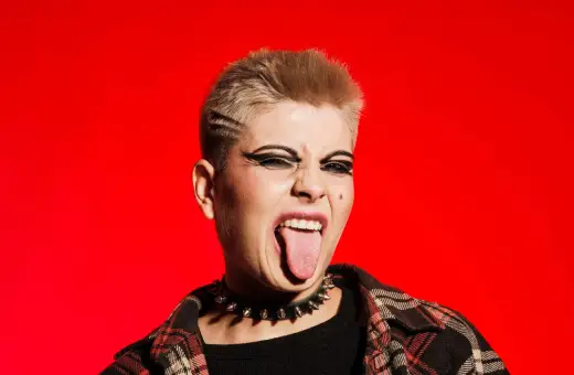 Młoda osoba z blond włosami i mocnym makijażem oczu, w kolczatce i flanelowej koszuli, wyciąga język. To kwintesencja buntu i stylu punk.