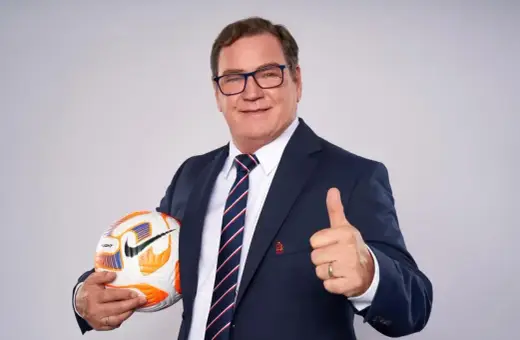 Uśmiechnięty trener reprezentacji Polski w garniturze trzyma piłkę i pokazuje kciuk w górę.