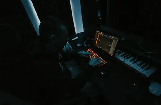 Producent muzyczny tworzy przebój w ciemnym studiu, pracując przy laptopie i klawiaturze sterującej.