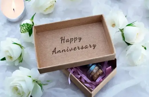 Otwarta szkatułka z napisem "Happy Anniversary" i małym prezentem w środku, otoczona białymi różami i płonącą świecą. Idealna na rocznice ślubu.