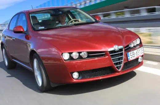 Czerwone Alfa Romeo 159 1.9 JTD pędzi po drodze, kierowca skupiony na trasie.