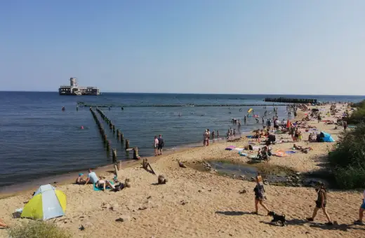 Słoneczna babie doły plaża tętni życiem. Ludzie opalają się, kąpią w morzu, a w oddali widać zabytkowy fort.