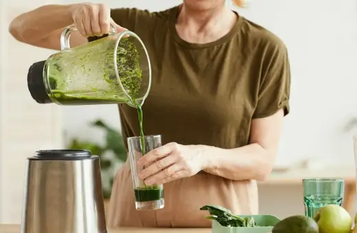 Kobieta nalewa zielony koktajl położnej do szklanki. Na blacie widać blender, awokado i jabłko.