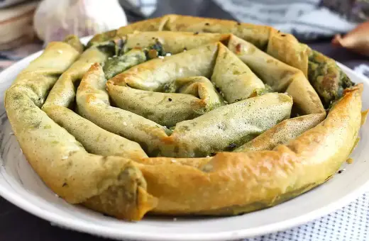 En gyllene, spiralformad burek med spenatfyllning, perfekt för ett hemlagat burek recept. Vitlök och lök i bakgrunden.