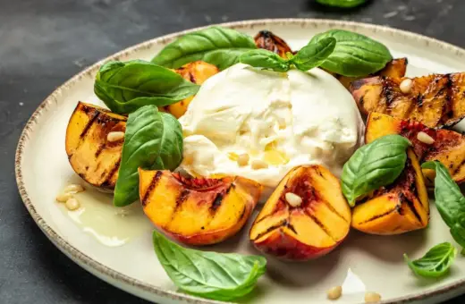 Burrata z grillowanymi brzoskwiniami, bazylią i orzeszkami piniowymi na talerzu.