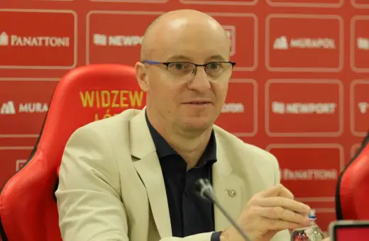 Nowy trener Widzewa w jasnej marynarce, siedzi na czerwonym fotelu z logo klubu.