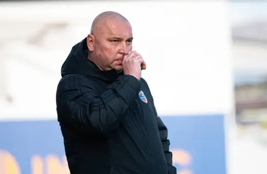 Mariusz Lewandowski, trener w czarnej kurtce, zamyślony, z ręką przy nosie.