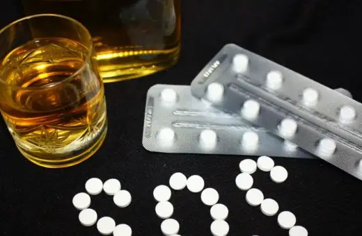 Szklanka alkoholu i blistry z tabletkami, ułożone w napis SOS. To ostrzeżenie przed niebezpiecznym połączeniem alkoholu i benzodiazepiny.