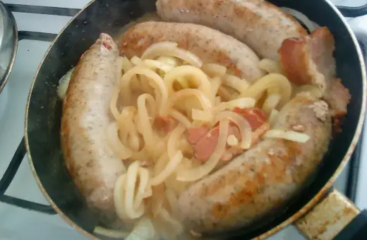 Biała kiełbasa w piwie dusi się z cebulą i boczkiem na patelni.