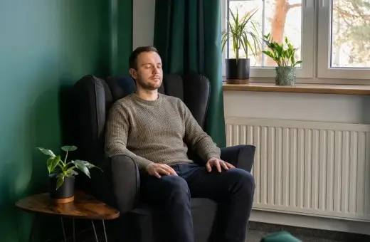 Mężczyzna w fotelu z zamkniętymi oczami, być może doświadcza objawów nerwicy. W tle zielona ściana, rośliny i okno.