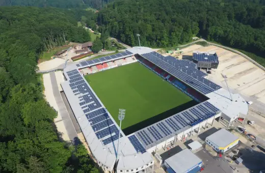 Widok z lotu ptaka na zieloną murawę i trybuny stadionu, otoczonego lasem. To najmniejszy stadion na świecie, z panelami słonecznymi na dachu.