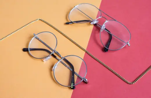 Dwa stylowe okulary, okrągłe i aviator, leżą na pomarańczowo-czerwonym tle. Złoty łańcuszek oplata je, sugerując, że to okulary korekcyjne, być może na astygmatyzm.