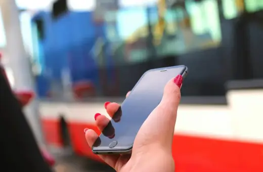 Dłoń z czerwonymi paznokciami trzyma smartfon, na którym można kupić bilety ZTM Warszawa. W tle widać autobus.