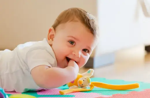 Uśmiechnięte 11 miesięczne dziecko leży na macie edukacyjnej, gryząc paluszek. Obok pluszowa zabawka.