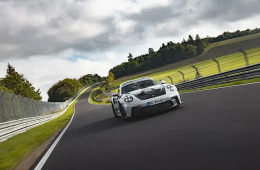 Biały Porsche 911 GT3 RS z czarnymi akcentami pędzi po torze wyścigowym wśród zielonych wzgórz i chmur.