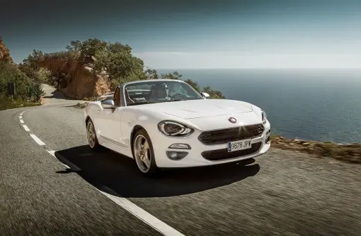 Biały Fiat 124 Spider sunie po krętej drodze nad morzem.