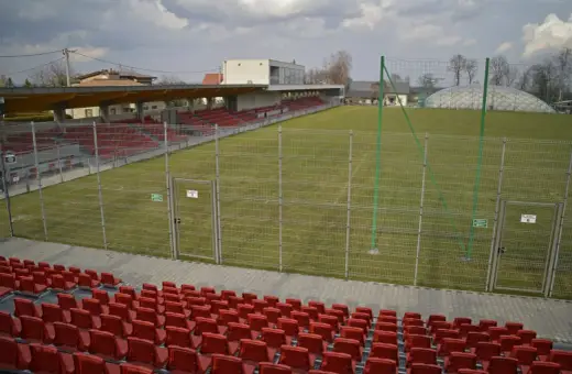 Puste trybuny i zielona murawa na stadionie w Niepołomicach. W tle budynek klubowy i balon nad boiskiem.