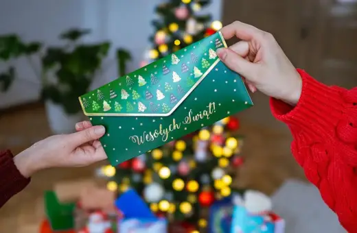 Jak zapakować voucher na prezent? Zielona koperta z choinkami i napisem "Wesołych Świąt!" jest przekazywana w świątecznej atmosferze.
