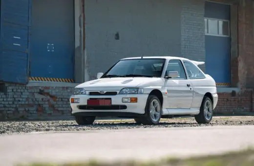 Biały Ford Escort Cosworth z charakterystycznym spojlerem, zaparkowany przed industrialnym budynkiem z niebieskimi drzwiami.